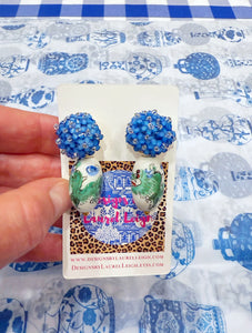 A hand is holding a pair of Chinoiserie earrings on a Designs by Laurel Leigh logo card. Earrings have blue hydrangeas and green dragons. Ginger jar print paper is in the background. 